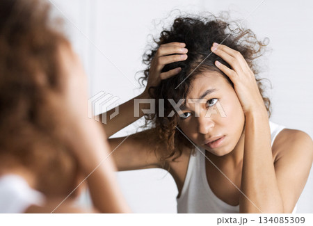 Afro-american girl frowning and looking intently on her face in mirror. Hair and sculp problems concept Afro-american girl frowning and looking intently on her face in mirror. Hair and sculp problems concept 134085309