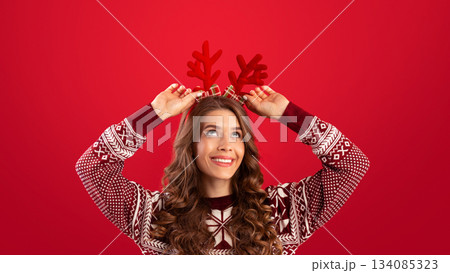 It's Christmas time. Pretty young woman in woolen sweater wearing deer horns on red studio background, panorama. Happy millennial lady having Xmas fun, celebrating New Year It's Christmas time. Pretty young woman in woolen sweater wearing deer horns on red studio background, panorama. Happy millennial lady having Xmas fun, celebrating New Year 134085323