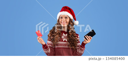 Online Christmas shopping. Lovely millennial girl in Santa hat and warm sweater holding mobile phone and credit card on blue background. Panorama Online Christmas shopping. Lovely millennial girl in Santa hat and warm sweater holding mobile phone and credit card on blue background. Panorama 134085400