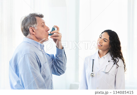 Medical Advice. Elderly man using asthma inhaler, mexican doctor watching, free space Medical Advice. Elderly man using asthma inhaler, mexican doctor watching, free space 134085415