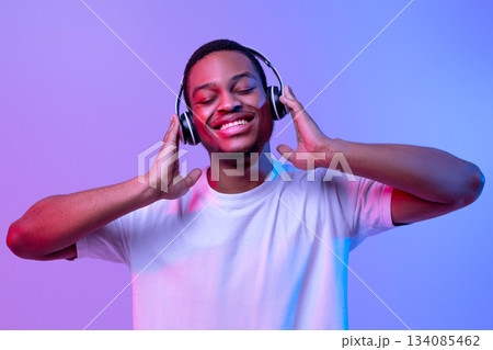 Music Streaming Services. Portraif Of Smiling Black Guy In Wireless Headphones Enjoying His Favorite Playlist, Standing With Closed Eyes Under Neon Lighting Over Purple Background, Panorama Music Streaming Services. Portraif Of Smiling Black Guy In Wireless Headphones Enjoying His Favorite Playlist, Standing With Closed Eyes Under Neon Lighting Over Purple Background, Panorama 134085462
