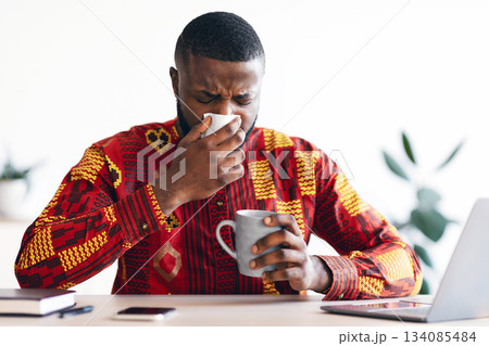 Sick Black Man In Ethnic African Shirt Sneezing At Workplace In Office, Blowing Nose To Napkin, Ill Millennial Man In Traditional Costume Suffering Seasonal Flu Or Having Coronavirus Symptoms 134085484