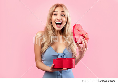 Excited blonde woman opening heart shaped gift box for Valentine's Day over pink studio background. Gorgeous millennial lady happy to receive present for lover's holiday 134085489