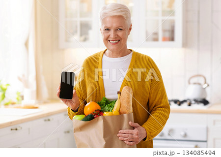 Cheerful senior woman with shopping bag full of grocery showing smartphone with blank screen, kitchen interior, copy space, mockup. Smiling old lady using mobile app for grocery delivery Cheerful senior woman with shopping bag full of grocery showing smartphone with blank screen, kitchen interior, copy space, mockup. Smiling old lady using mobile app for grocery delivery 134085494