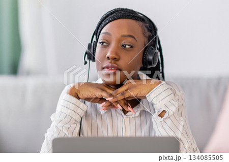 Pensive young black lady having online training at home, sitting at workdesk in front of laptop, leaning on her elbows and looking at copy space, using headphones. E-education, online class concept Pensive young black lady having online training at home, sitting at workdesk in front of laptop, leaning on her elbows and looking at copy space, using headphones. E-education, online class concept 134085505