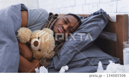 Depressed Sick African Man Hugging Teddy Bear Toy Lying Wrapped In Blanket Having Cold In Bedroom At Home. Flu And Depression, Mental Health During Coronavirus Disease Concept. Selective Focus 134085506