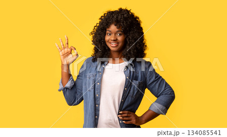 Happy beautiful black woman with curly hair standing on yellow studio background, showing okay gesture, copy space. Happy and carefree african american young lady in casual jeans shirt enjoying life 134085541