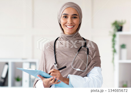 Cheerful pretty woman in hijab doctor posing in cabinet, holding medical folder and pen, having stethoscope, copy space. Happy muslim lady greeting patients at hospital, smiling at camera 134085672