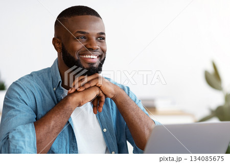 Portrait of pensive smiling black freelancer sitting at desk in home office, thinking about business strategy while working on laptop computer, looking away with pleased face expression, free space Portrait of pensive smiling black freelancer sitting at desk in home office, thinking about business strategy while working on laptop computer, looking away with pleased face expression, free space 134085675