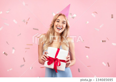 Happy young lady in birthday hat and cute dress holding gift box on pink studio background with falling confetti. Attractive millennial blonde celebrating holiday, having fun party Happy young lady in birthday hat and cute dress holding gift box on pink studio background with falling confetti. Attractive millennial blonde celebrating holiday, having fun party 134085695