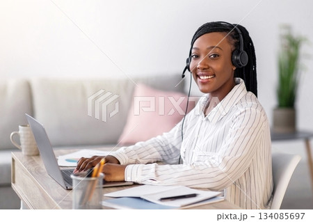 Pretty black woman smiling at camera while listening to webinar from home, using headset and laptop, side view, copy space. Cheerful african american lady working online, typing on computer keyboard 134085697