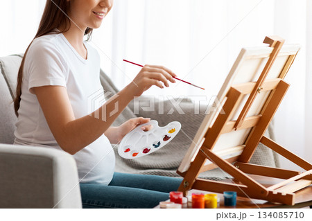 Pregnancy Leisure. Happy pregnant woman painting at home, sitting on couch near easel, holding color palette, smiling expectant mother enjoying drawing art therapy, side view with copy space 134085710