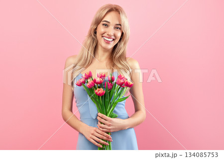 Portrait of romantic blonde woman in dress smiling and holding spring tulips on pink studio background. Joyful young lady posing with lovely flowers. Holiday celebration concept Portrait of romantic blonde woman in dress smiling and holding spring tulips on pink studio background. Joyful young lady posing with lovely flowers. Holiday celebration concept 134085753