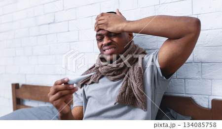 Fever And Flu. Ill Black Guy Touching Forehead Holding Thermometer Having Influenza Symptoms And Headache Sitting In Bed Indoors. Covid-19 Virus, Seasonal Health Problems. Selective Focus 134085758