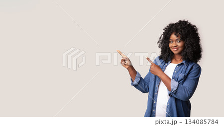 Positive black curly woman pointing at copy space over gray studio background, panorama. Young pretty african american lady smiling and indicating to empty space for text or advertisement Positive black curly woman pointing at copy space over gray studio background, panorama. Young pretty african american lady smiling and indicating to empty space for text or advertisement 134085784