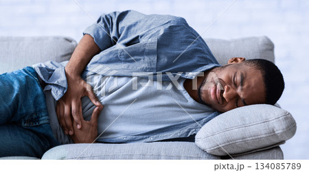 Stomachache. African Guy Suffering From Abdominal Pain Touching Aching Stomach Lying On Sofa At Home. Black Man Having Gastritis Problem And Abdomen Inflammation. Health Issue Concept 134085789