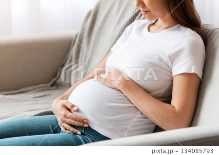 Prenatal Time. Beautiful Future Mother Sitting On Couch Embracing Her Belly, Happy Pregnant Woman Relaxing on Sofa In Living Room At Home, Tenderly Touching Tummy And Smiling, Side View, Free Space Prenatal Time. Beautiful Future Mother Sitting On Couch Embracing Her Belly, Happy Pregnant Woman Relaxing on Sofa In Living Room At Home, Tenderly Touching Tummy And Smiling, Side View, Free Space 134085793
