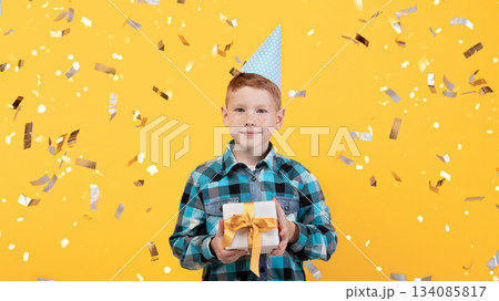 Cute little redhead boy celebrating birthday over yellow studio background, wearing festive paper cap, holding gift box, throwing confetti. Happy kid having birthday party, posing alone, studio shot 134085817