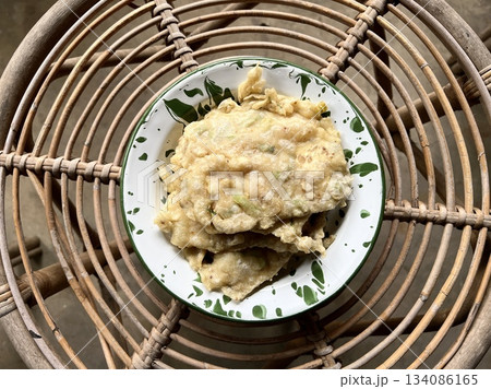 Tempe Mendoan, an Indonesian traditional snack made from tempe or tempeh (fermented soybeans) coated in flour and spring onions and fried half-cooked, on a white enamel plate, placed on a rattan table 134086165