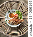 Fried tilapia served on a white enamel plate with a green rim, accompanied by chili sauce, and fresh vegetables specifically lettuce, sliced cucumber, and tomato. Placed on a rattan woven table. 134086166