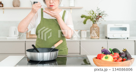 マイホームのキッチンで料理中に味見をするエプロン姿のシニア女性 134086806