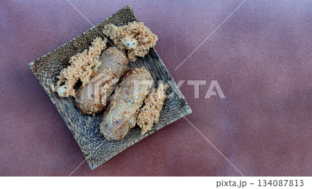 Above view of Khao Mao dessert or name Pounded Unripe Rice or Shredded Rice Grain. Dish made from ripe bananas wrapped in rice and fried. Placed on a wooden plate on a light orange tiled floor. Above view of Khao Mao dessert or name Pounded Unripe Rice or Shredded Rice Grain. Dish made from ripe bananas wrapped in rice and fried. Placed on a wooden plate on a light orange tiled floor. 134087813