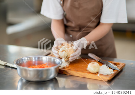 Close up of gloved hands chef cleaning white onion peels or skins on wooden chopping board. Close up of gloved hands chef cleaning white onion peels or skins on wooden chopping board. 134089241