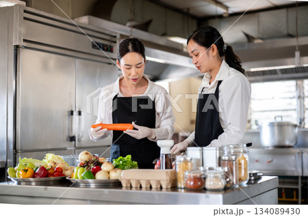 Female chef hold carrot while teaching assistant or student at kitchen counter with food ingredients Female chef hold carrot while teaching assistant or student at kitchen counter with food ingredients 134089430