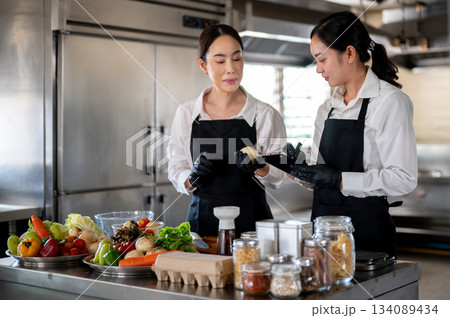 Female chef teaching a student or assistant in kitchen at cooking counter full of food ingredients. 134089434