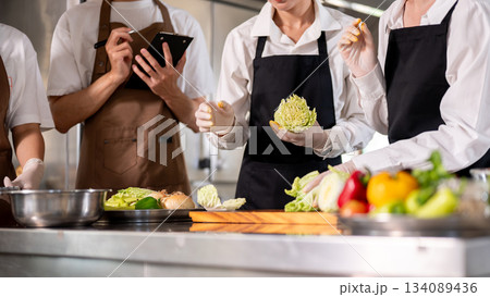 Close up of chef holding cabbage cuts while standing with students or assistants at kitchen counter. Close up of chef holding cabbage cuts while standing with students or assistants at kitchen counter. 134089436