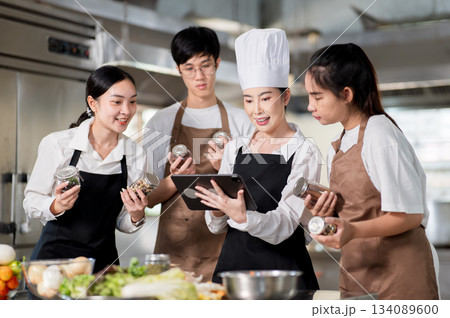 Asian female chef holding tablet showing cooking recipe to her students with seasonal jars in hands. Asian female chef holding tablet showing cooking recipe to her students with seasonal jars in hands. 134089600