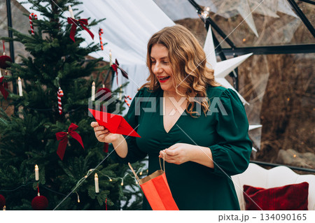 Young 30s woman getting Christmas greeting card from her beloved on background of xmas tree at cozy living room with festive interior. Surprised lady pleasure reading letter at home. 134090165