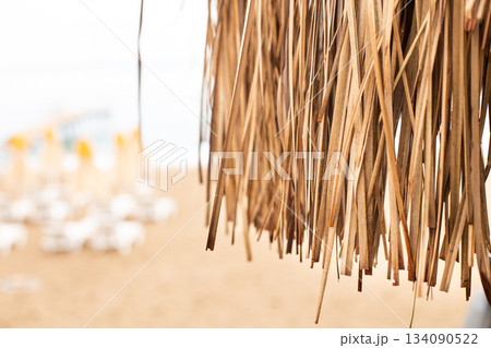 Thatched roof with tropical beach with sand and deck chairs on the sea shore in background. 134090522