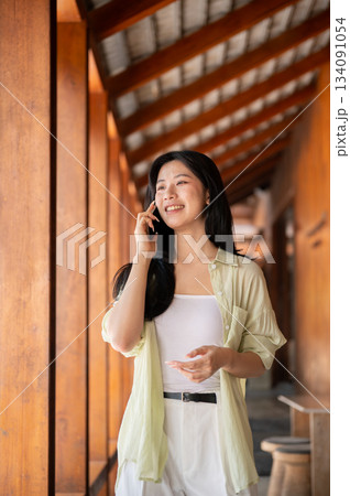 Long hair asian woman talking on a phone and looking ahead while walking in a wooden house's corridor. 134091054