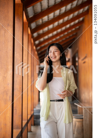 Long hair asian woman is smiling while talking on a smartphone in a wooden house's corridor. Long hair asian woman is smiling while talking on a smartphone in a wooden house's corridor. 134091055