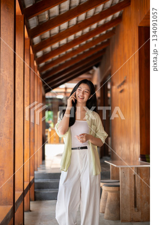 Long hair asian woman is standing and talking on a smartphone in the wooden house's corridor. Long hair asian woman is standing and talking on a smartphone in the wooden house's corridor. 134091057