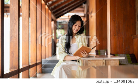 Long hair asian woman is reading a book while sitting at table in wooden house's corridor. 134091068
