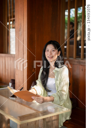 Asian woman is smiling with a book and a pen in her hands while sitting at table in a wooden house. Asian woman is smiling with a book and a pen in her hands while sitting at table in a wooden house. 134091076