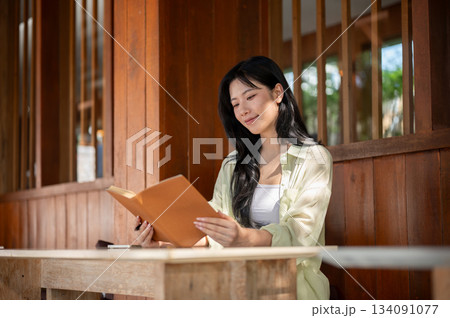 Long hair asian woman is smiling and reading a book while sitting at table in a wooden house. Long hair asian woman is smiling and reading a book while sitting at table in a wooden house. 134091077
