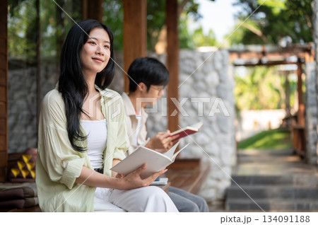 Long hair asian woman looking away from a book in hand while sitting next to a man at wooden terrace 134091188