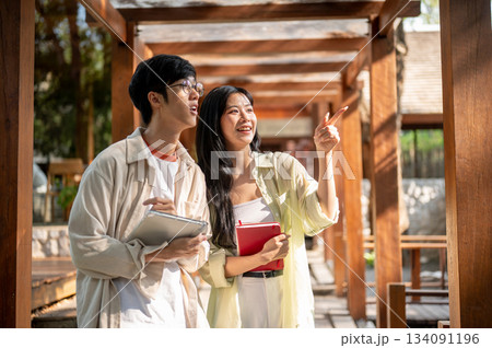 Asian woman pointing a finger showing to her male friend while standing in a wooden corridor. Asian woman pointing a finger showing to her male friend while standing in a wooden corridor. 134091196