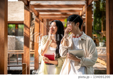 Smiling asian woman and a male friend standing with a hand covered mouth in garden wooden corridor. Smiling asian woman and a male friend standing with a hand covered mouth in garden wooden corridor. 134091200