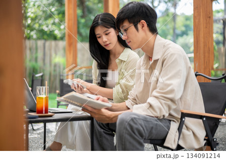 Asian man with glasses holding notebook and reading together with female friend in garden cafe. Asian man with glasses holding notebook and reading together with female friend in garden cafe. 134091214