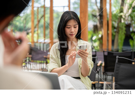 Long hair asian woman is holding a smartphone reading chats while sitting on folding chair in cafe. Long hair asian woman is holding a smartphone reading chats while sitting on folding chair in cafe. 134091231