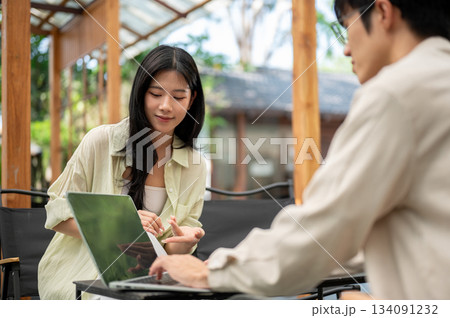 Smiling asian woman leaning to look at friend's laptop screen while sitting on folding chair in cafe Smiling asian woman leaning to look at friend's laptop screen while sitting on folding chair in cafe 134091232