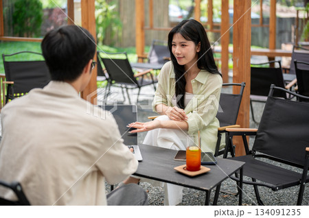 Asian woman smiling and pointing at laptop screen to a friend while sitting on folding chair in cafe 134091235