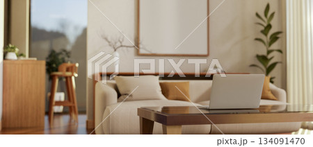 White Laptop on a glass top wooden table next to fabric sofa in a room with blank frame on the wall. 134091470