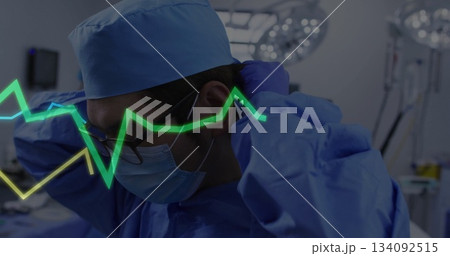 Mature surgeon in blue scrubs adjusting gloves, cap under surgical lights in OR, with ECG overlay 134092515