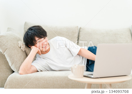 家のリビングでソファーに寝転びながらパソコンで動画視聴をする若いアジア人男性 家のリビングでソファーに寝転びながらパソコンで動画視聴をする若いアジア人男性 134093708