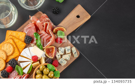 Top view of gourmet charcuterie and cheese board with prosciutto, brie, blue cheese, crackers, figs and berries on dark background with copy space 134093735